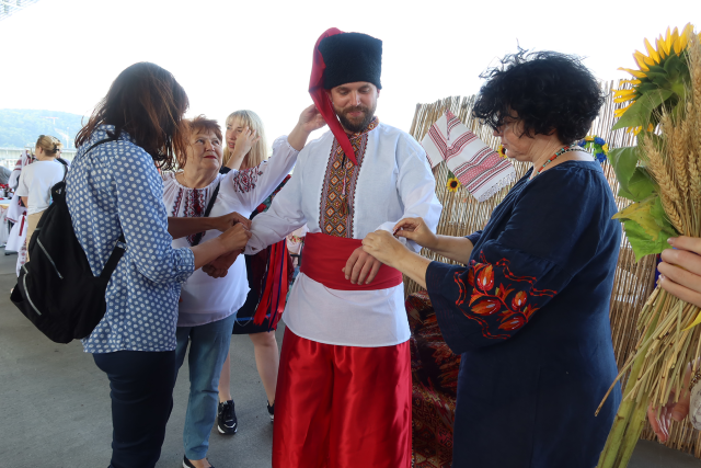Ein Mann ist von einigen Frauen umringt. Sie helfen ihm gerade dabei ukrainische Tracht anzuziehen. Er trägt ein weißes Hemd mit Stickereien, eine rote Hose und über dem Hemd einen roten Gürtel. Auf dem Kopf trägt er eine Stoffmütze mit schwarzer Krempe und rotem Zipfel