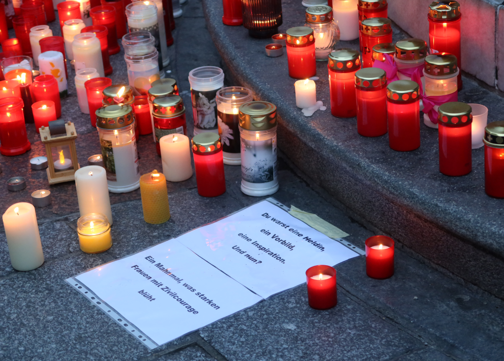 Vor einem Springbrunnen stehen viele brennende Kerzen und zwei Zettel mit folgendem Text: "Du warst eine Heldin, ein Vorbild, eine Inspiration. Und nun? Ein Mahnmal, was starken Frauen mit Zivilcourage blüht."