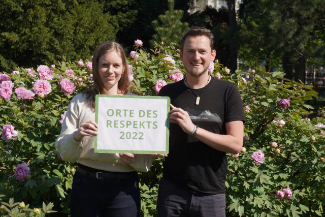 2 Personen, eine Frau und ein Mann, stehen vor einem Rosenbusch. Sie halten ein Schild hoch, auf dem steht: "Orte des Respekts 2022"