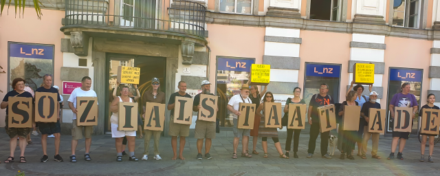 Personen, die in einer Reihe stehen, halten Schachteln vor sich, auf denen mit schwarzen Buchstaben die Wörter "Sozialstaat Adé" buchstabiert werden.