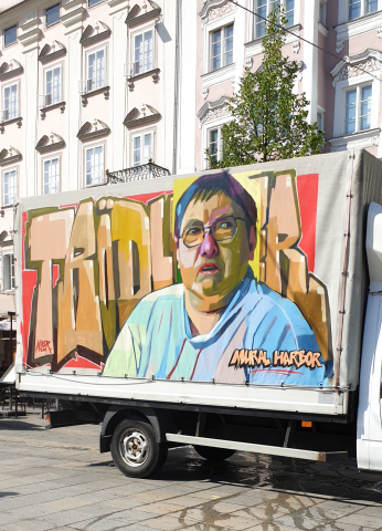 Auf der Seite eines weißen Lasters ist ein Portrait im Graffitistyle gesprayed. Es zeigt einen älteren Mann mit Brille, Hemd und grauen Haaren. Er blickt zur Seite. Im Hintergrund ist das Wort Trödler gesprayed in großen, dicken Buchstaben