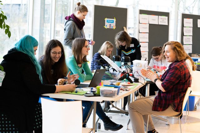 Junge Menschen sitzen an einem langen Tisch und haben Laptops vor sich. Sie arbeiten konzentriert, aber lachen auch miteinandern