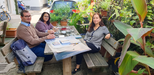 3 Personen sitzen auf einer Holzgarnitur in einem Innenhof. Sie schauen in die Kamera. Auf dem Tisch liegen Poster, Stifte und Wassergläser