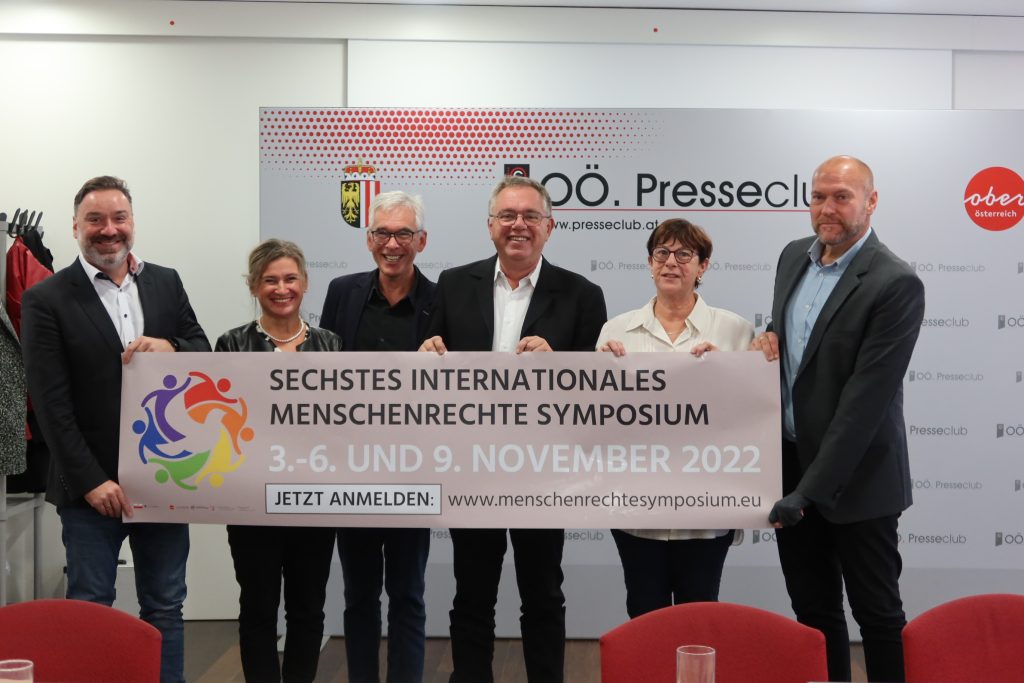 6 Menschen schauen in die Kamera und halten ein Banner hoch. Darauf ist zu lesen: "Sechstes Internationales Menschrechtesymposium. 3.-6. und 9. November 2022