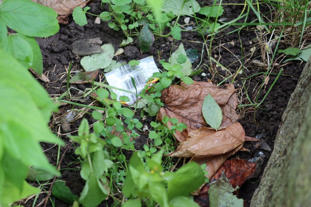 Auf einem erdigen Boden, zwischen grünen Sträuchern, liegt Plastikmüll. Es sieht wie eine Verpackung aus, auf der ein Strichcode zu sehen ist.