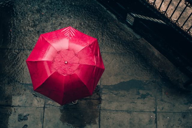 Aus der Vogelperspektive sieht man einen roten Regenschirm, von der Person, die ihn hält ist fast nichts zu sehen. Der Boden ist grau und nass.