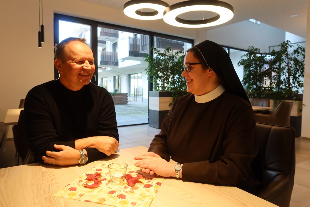 Zwei Menschen sitzen an einem Tisch und lachen sich an. Sie werden von oben angeleuchtet. Auf dem Tisch ist herbstliche Deko mit Blättern und Kerzen