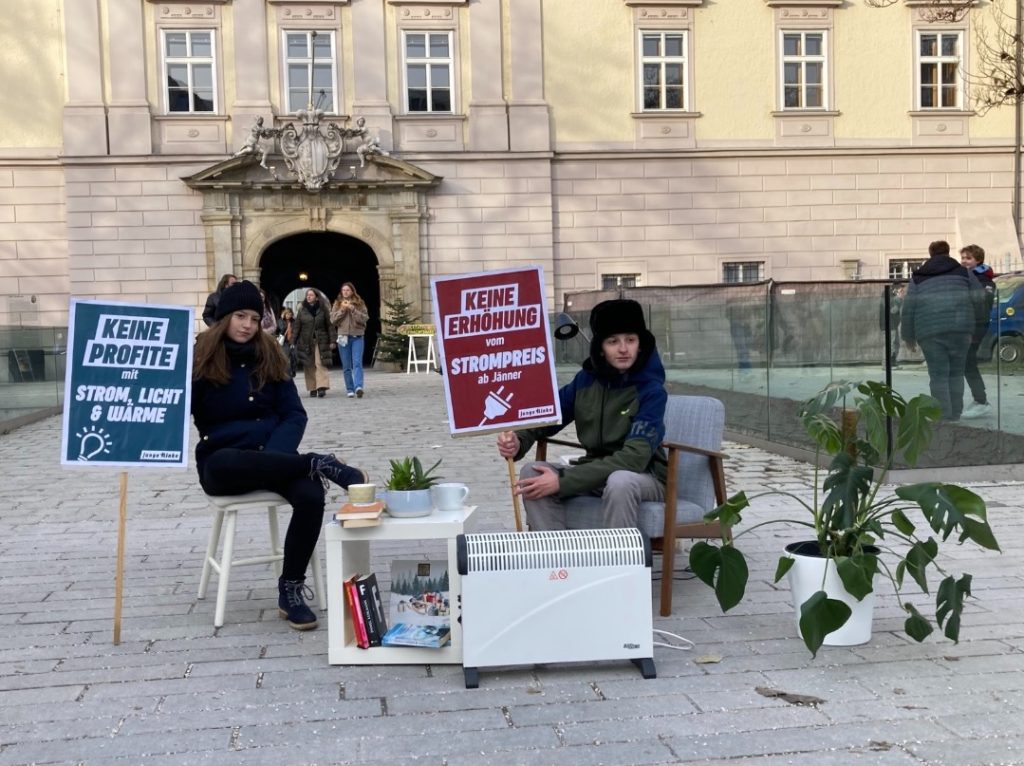 2 Personen sitzen draußen auf Sesseln. Sie sind warm angezogen. Vor ihnen steht ein Heizstrahler. Sie halten Plakate mit Aufschriften.