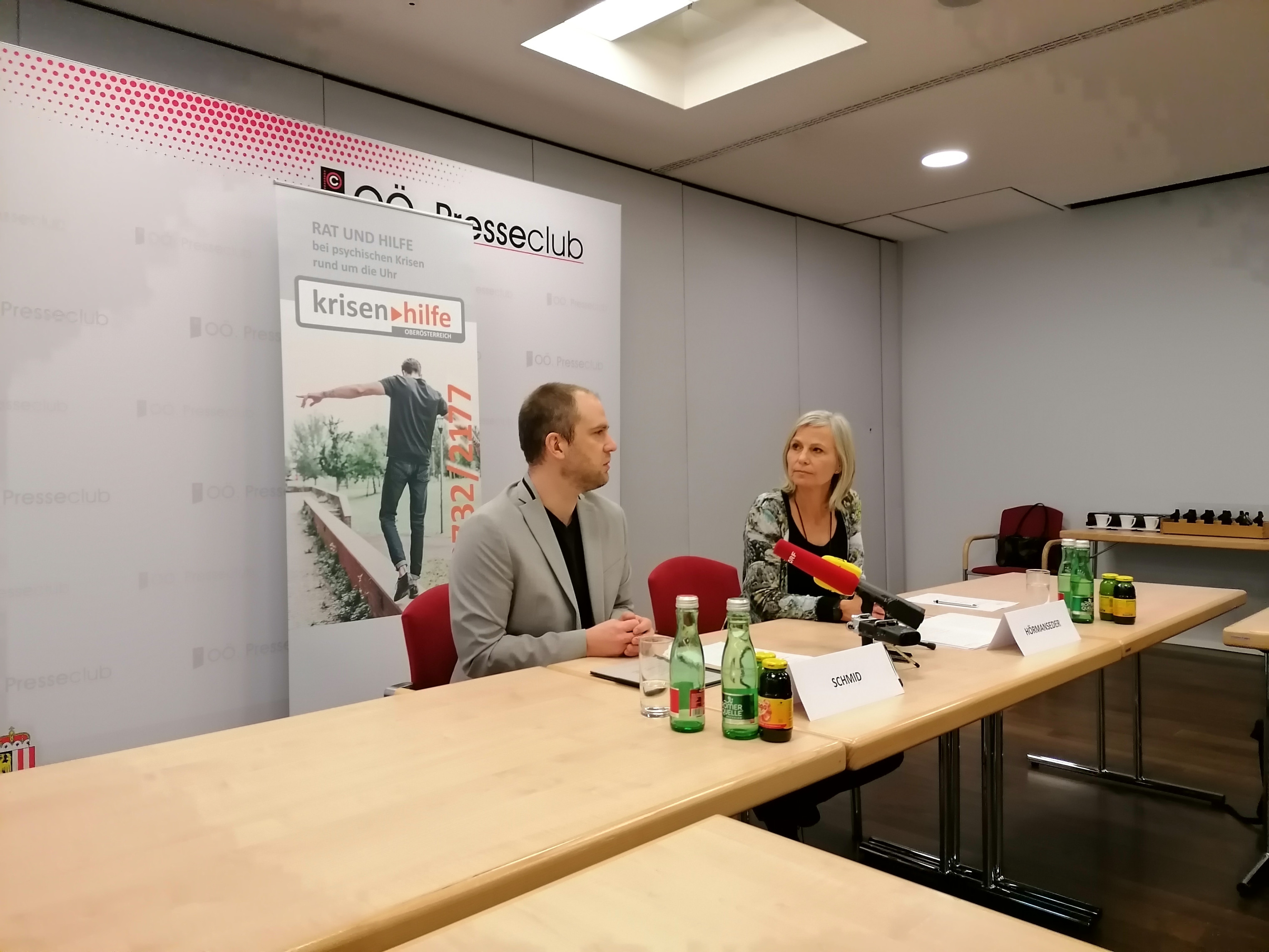 Zwei Menschen sitzen an einem Tisch und haben Mikrofone vor sich stehen. Es ist eine Pressekonferenz der Krisenhilfe.