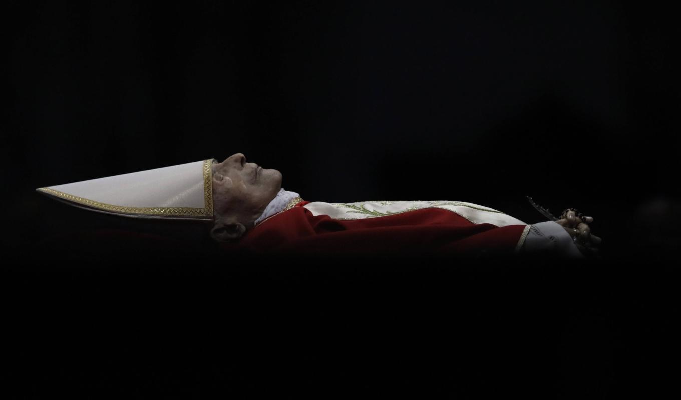 Der tote Papst Benedikt XVI liegt auf dem Rücken. Er ist von der Seite zu sehen. Rund um ihn herum ist es schwarz. Er trägt rot-weißes Gewand