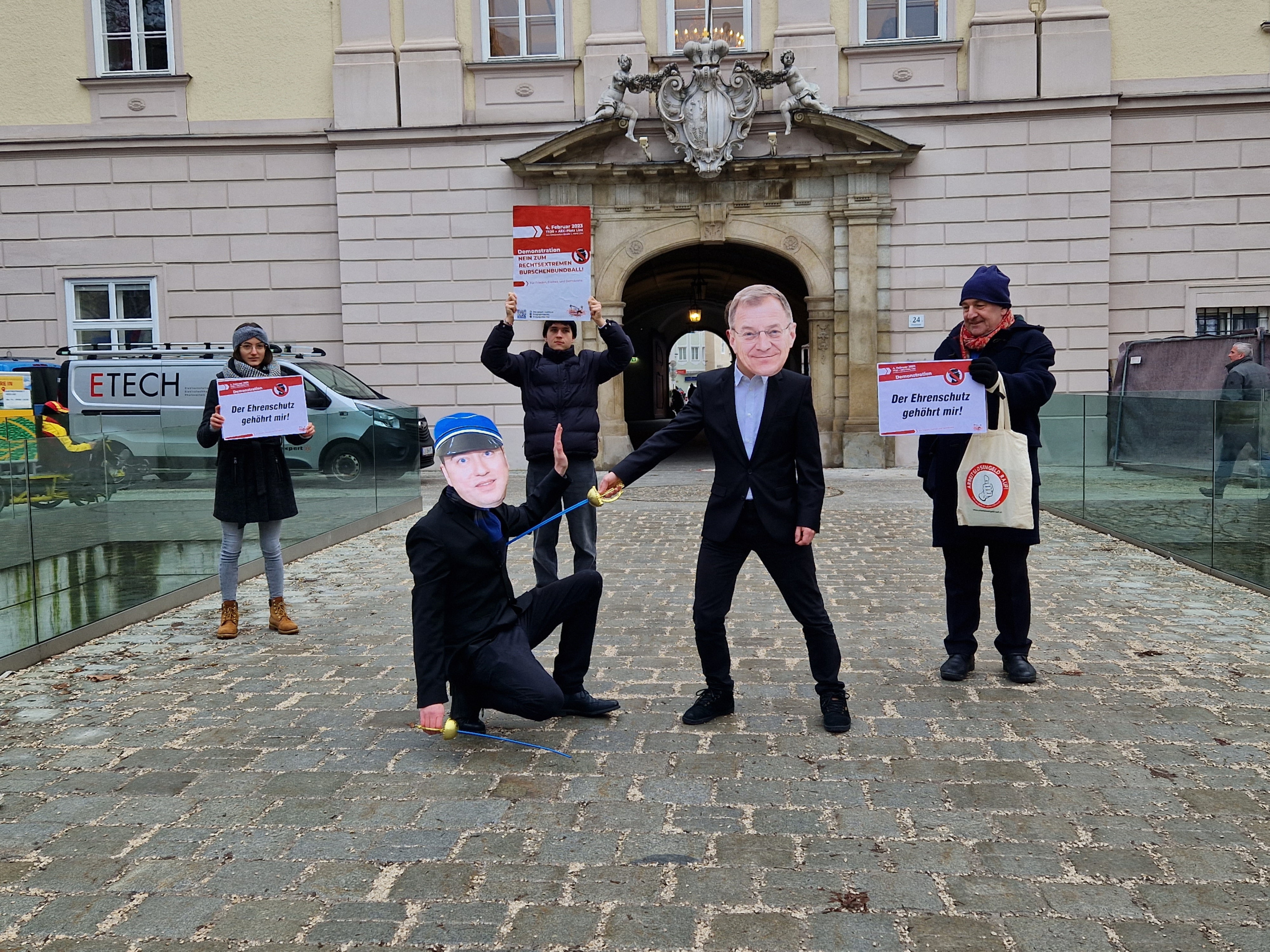 Personen, die sich als Thomas Stelzer und Manfred Haimbuchner verkleidet haben, haben ein simuliertes Säbelduell gekämpft. Stelzer hat gewonnen und steht rechts im Bild. Er hält sein Säbel an Haimbuchners Hals, der links neben ihm kniet.