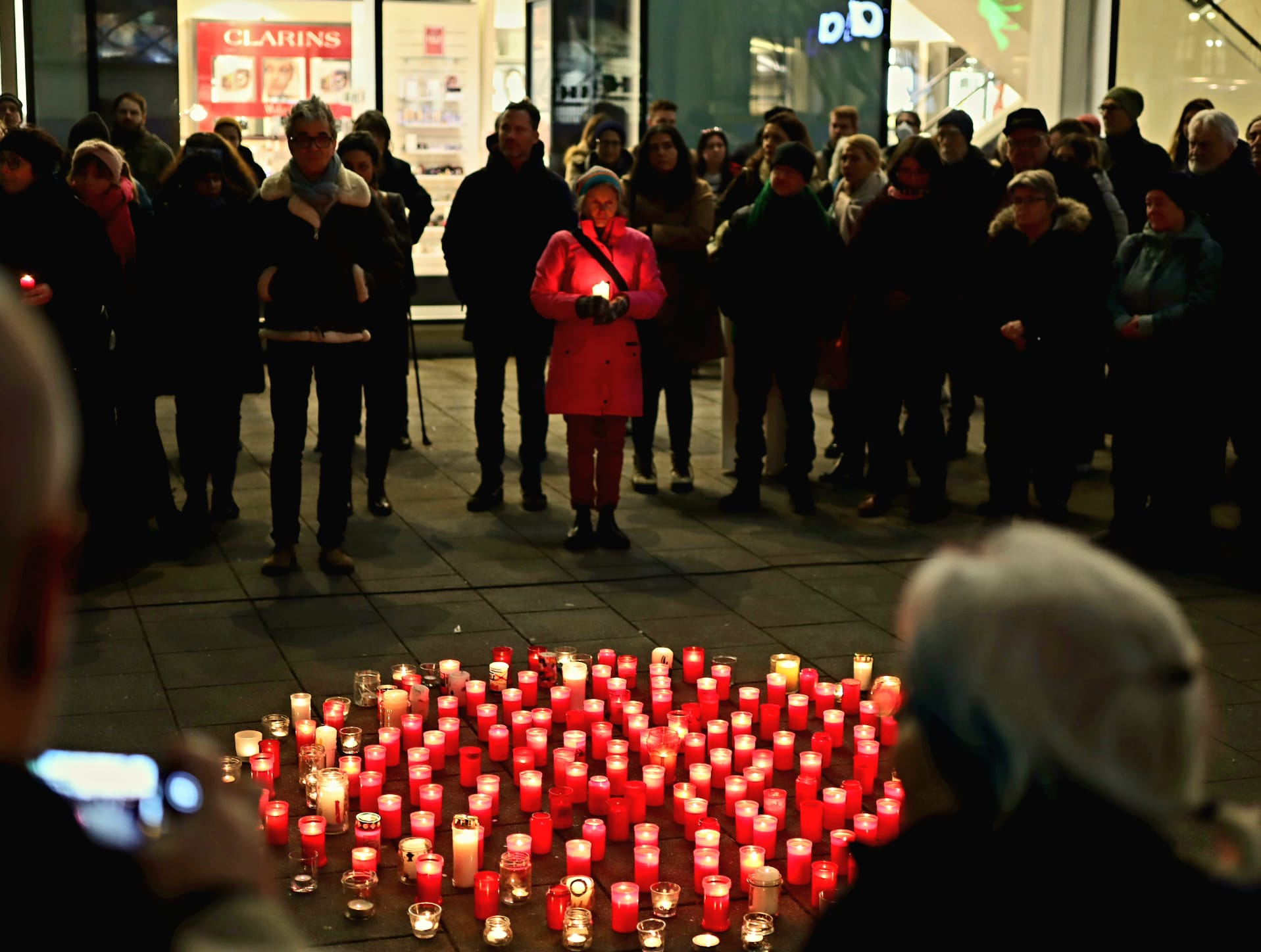 Am Boden stehen ganz viele rote Kerzen. Sie bilden einen Kreis. Es ist dunkel und man sieht die Menschen, die rundherum stehen nur schemenhaft. Eine Person hält eine Kerze in der Hand und ist heller erleuchtet.