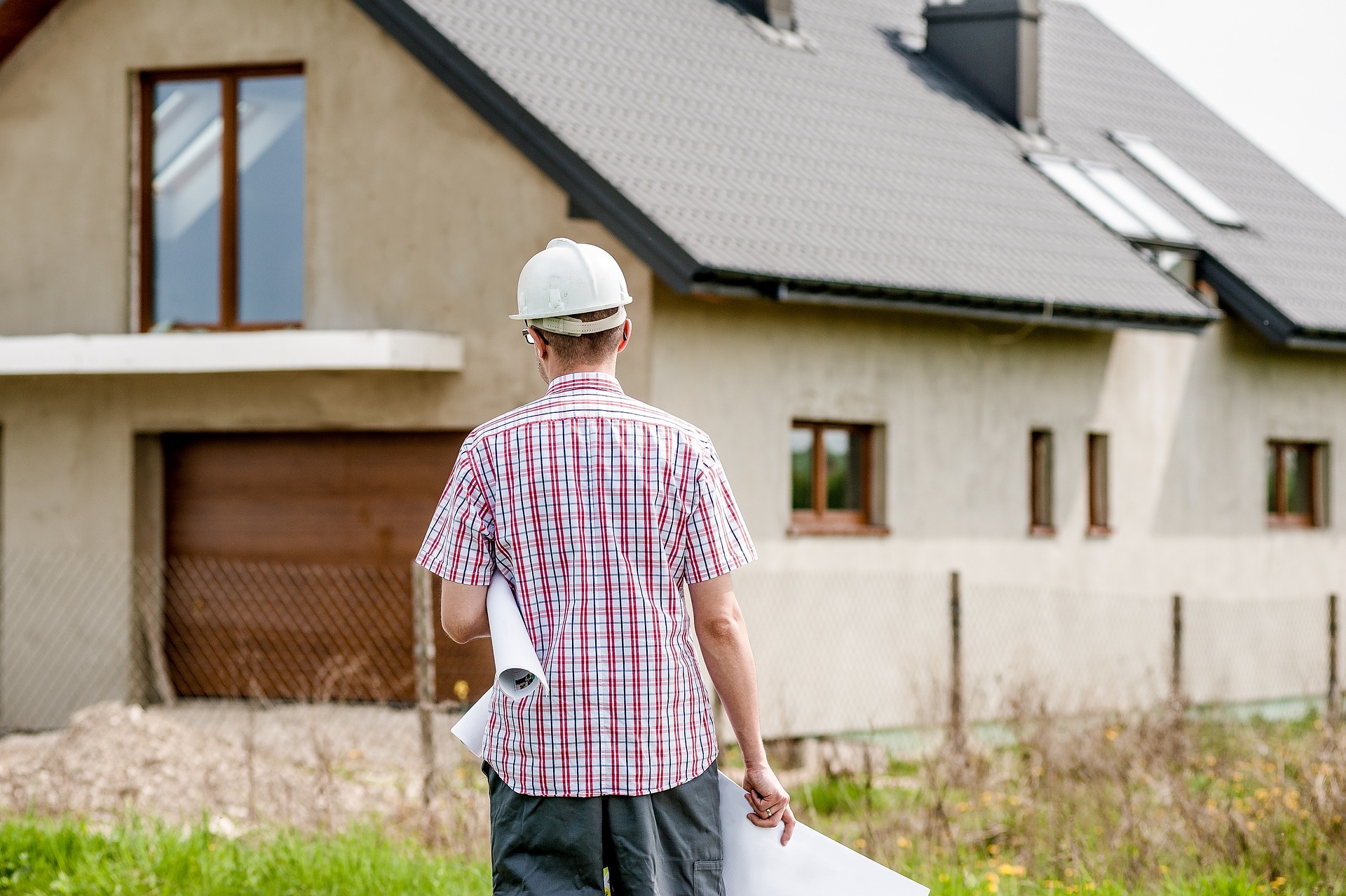 Ein Mann ist von hinten vor einem Haus zu sehen. Er trägt einen Helm für eine Baustelle und Pläne in der Hand.