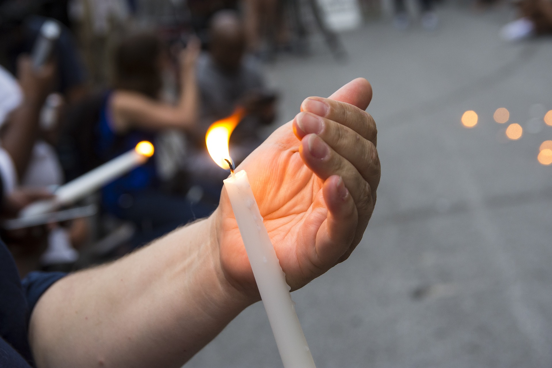 Eine Hand schützt eine Kerze vor dem Wind, damit eine andere Person die eigene Kerze an dieser entzünden kann.