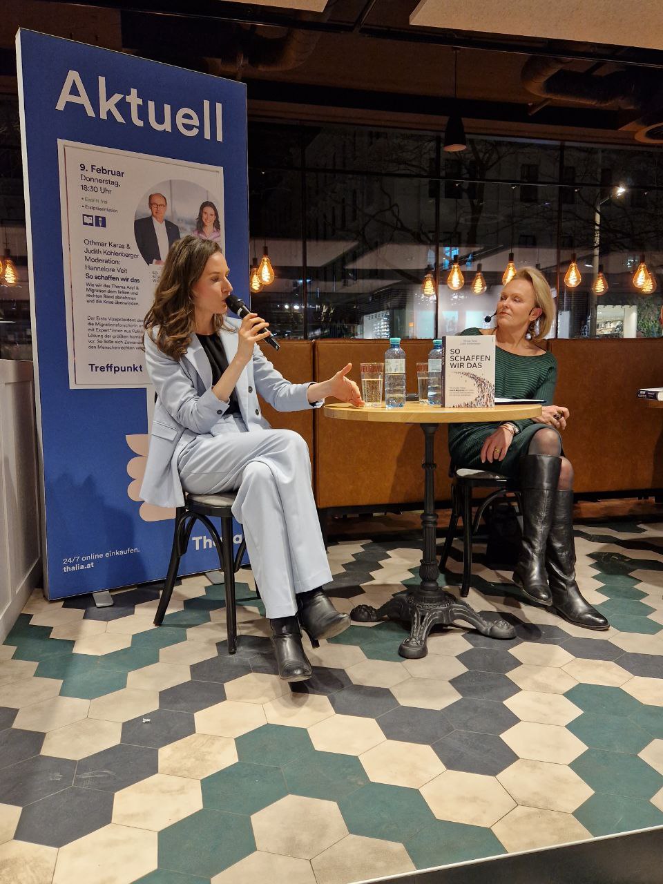 2 Frauen sitzen an einem runden Tisch nebeneinander. Sie halten eine Podiumsdiskussion und haben Mikrofone in der Hand.