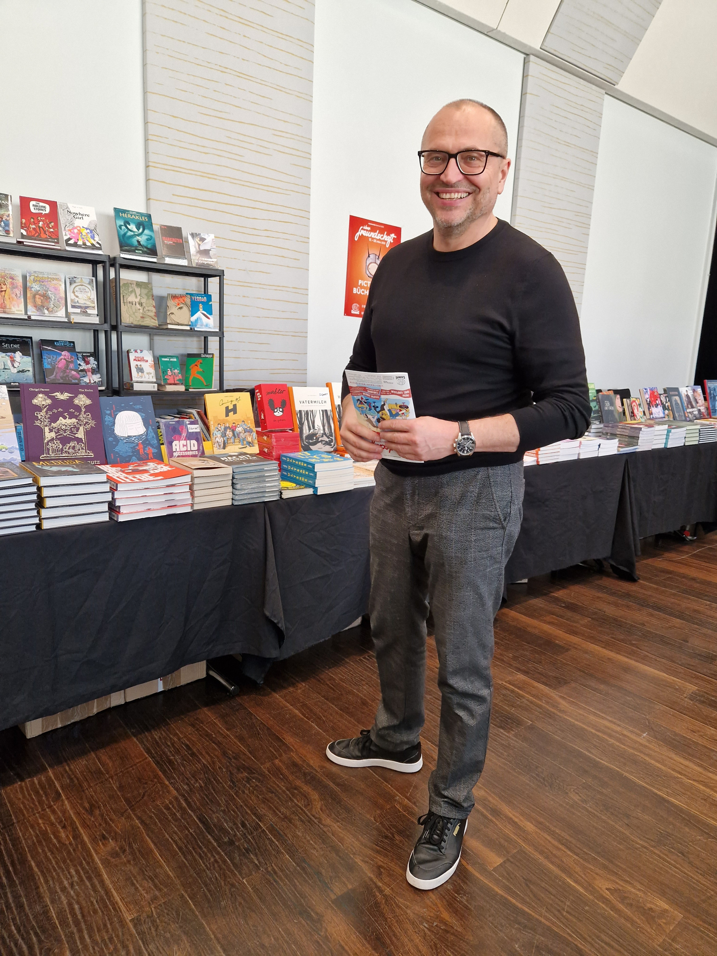 Ein Mann steht vor einem Tisch mit Comicbüchern und lächelt in die Kamera