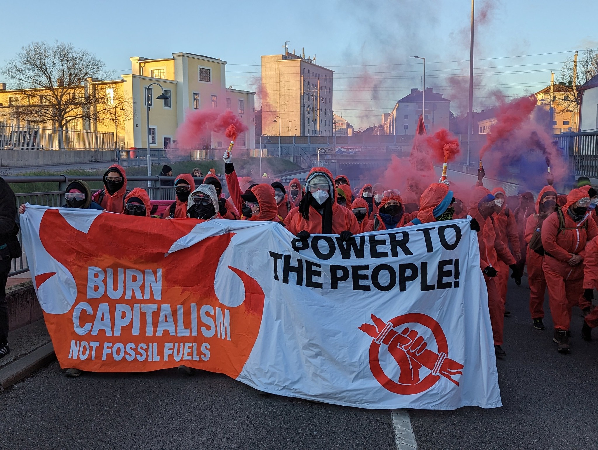 Eine Menge, in roten Overalls gekleideter Protestierender halten ein Banner hoch. Darauch steht: Burn capitalism, not fossil fuels. Power to the people
