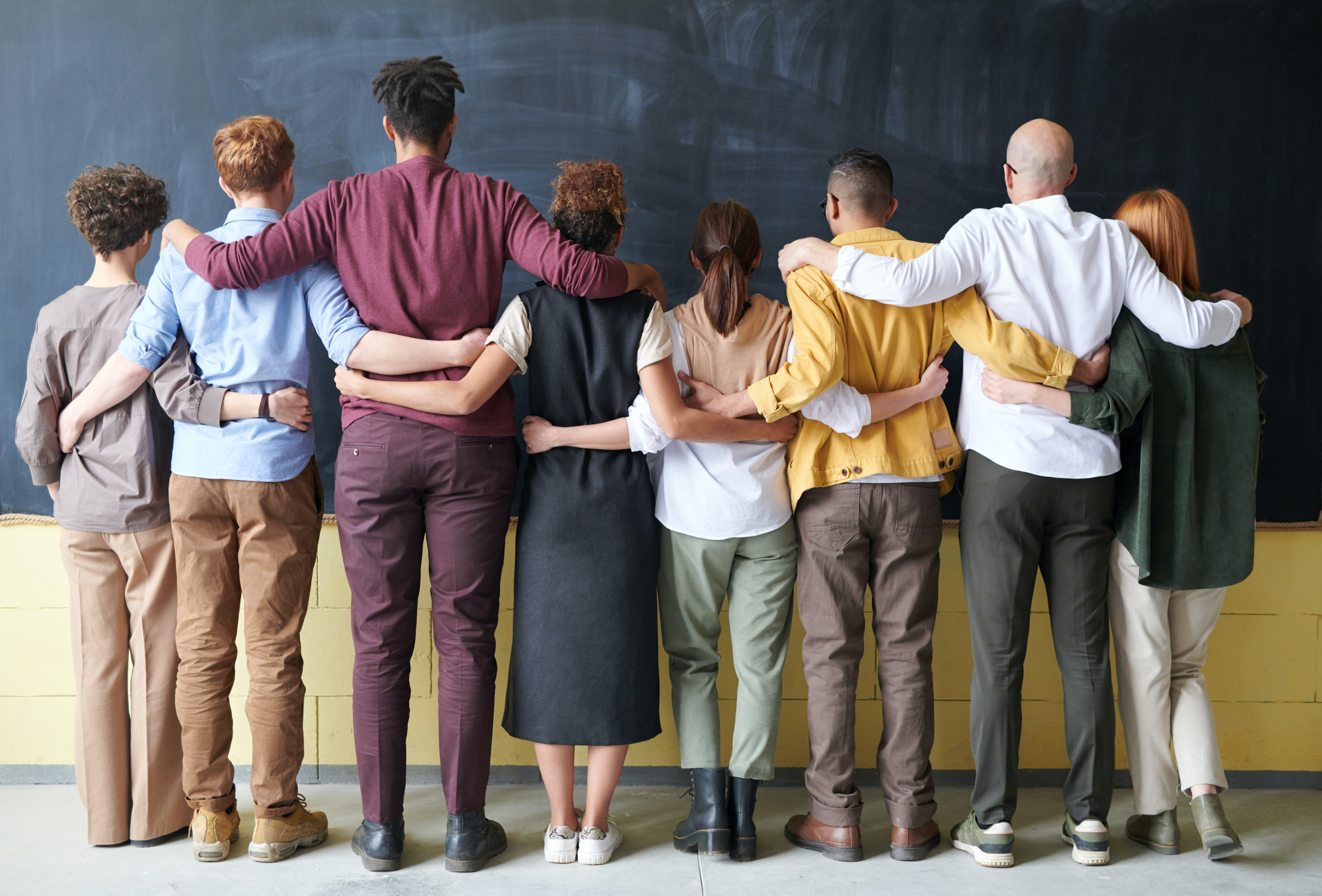 Man sieht eine Reihe von Menschen von hinten. Sie stehen vor einer Wandtafel und halten sich an Hüfte und Schulter von der Person neben sich fest