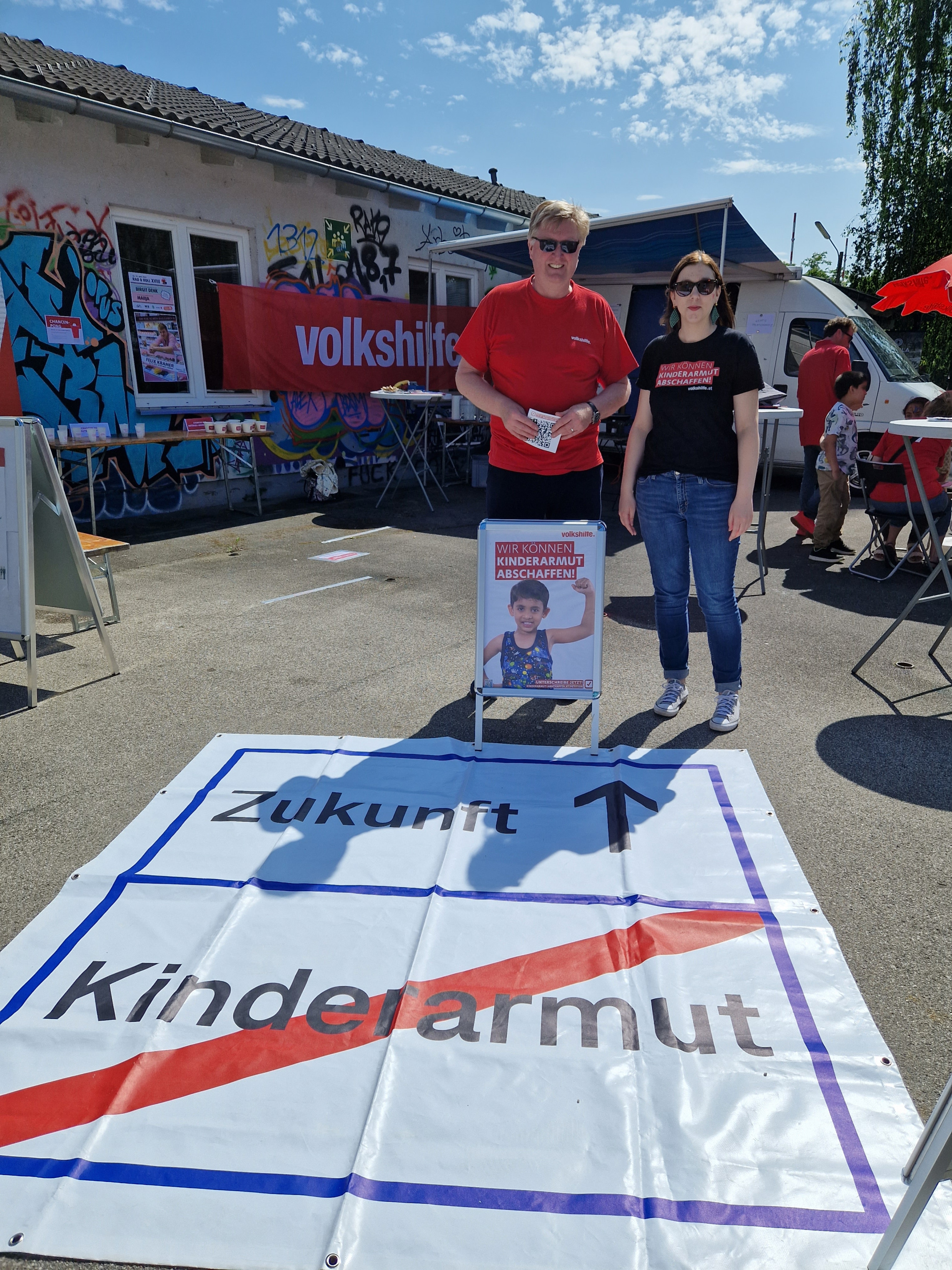 2 Personen stehen hinter einem Banner, das am Boden liegt. Auf diesem Banner ist zu lesen: Zukunft mit einem Pfeil nach oben und darunter ist durchgestrichen: Kinderarmut. Das Banner sieht aus wie ein Ortschild. Weiße Fläche mit blauem Rahmen