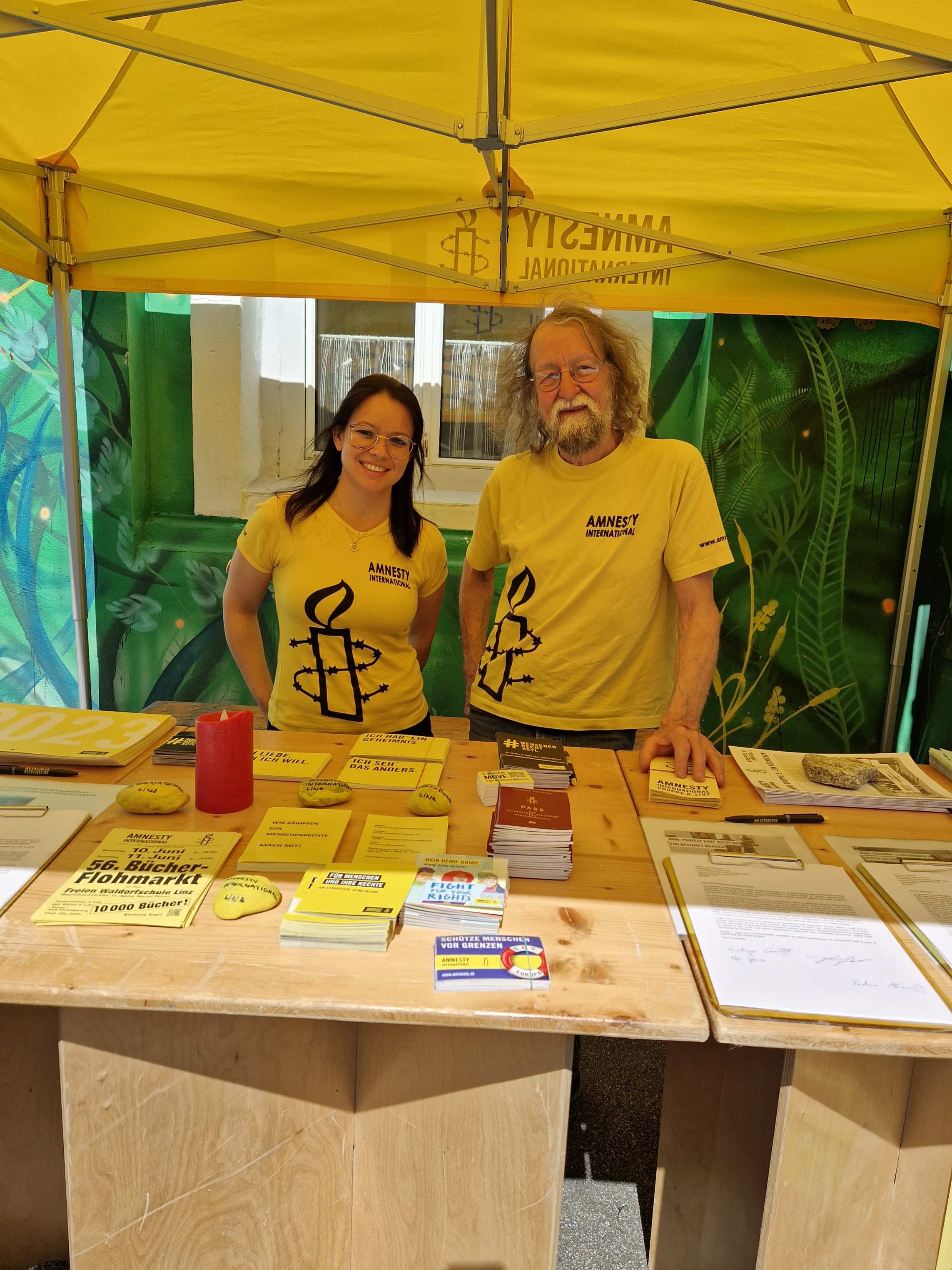 2 Personen stehen hinter einem Tisch mit Flyern und Informationszeitungen von Amnesty International. Sie tragen beide gelbe Tshirts.