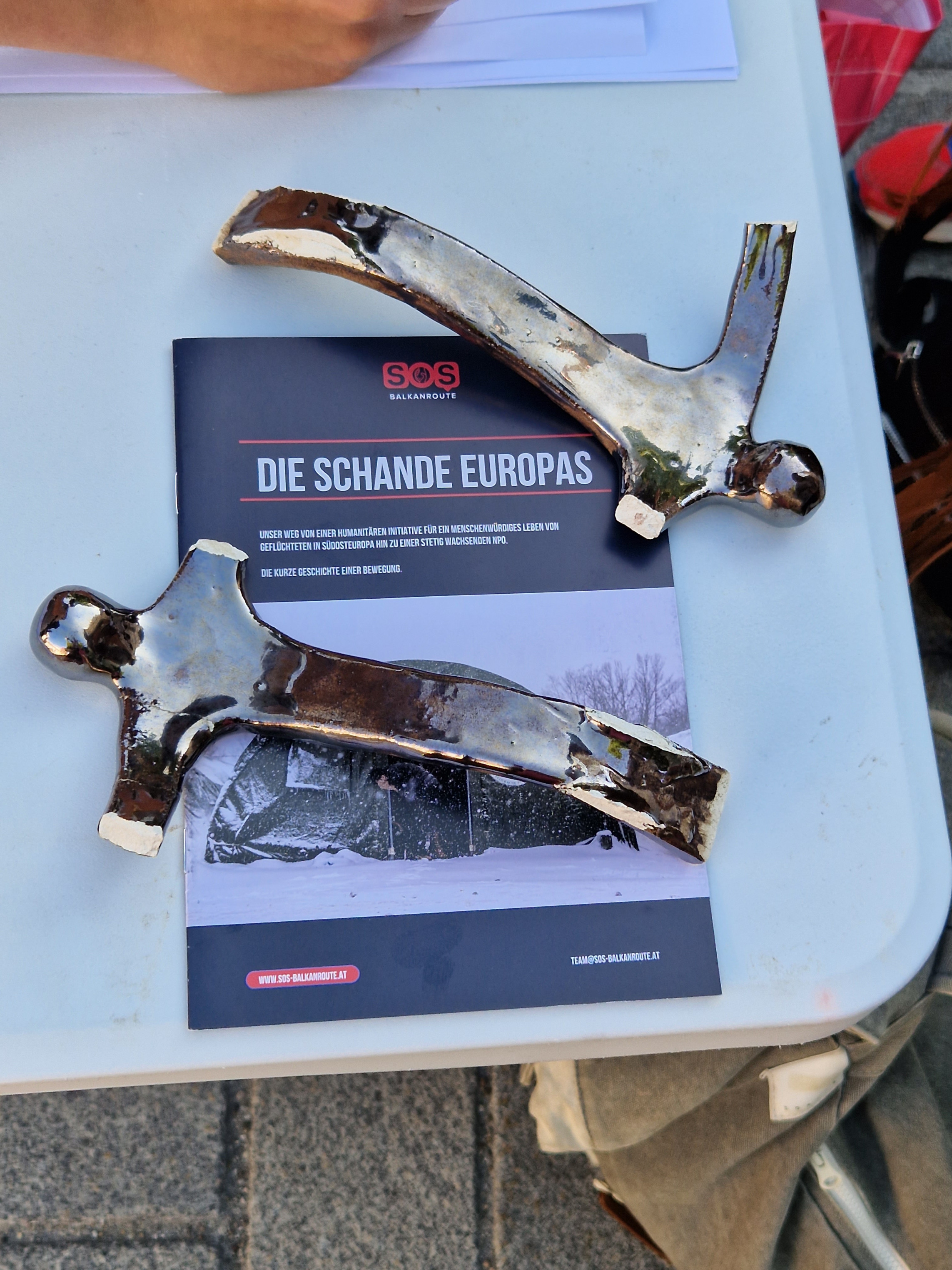 Zerbrochene Teile einer Statue liegen auf einem Flyer auf dem "Die Schande Europas" zu lesen ist. Die Teile sehen menschlich aus, die Farbe ist silber und es ist nur eine Silhoutte von Menschen zu sehen, ohne Mimik oder detaillierte Arme und Beine. 