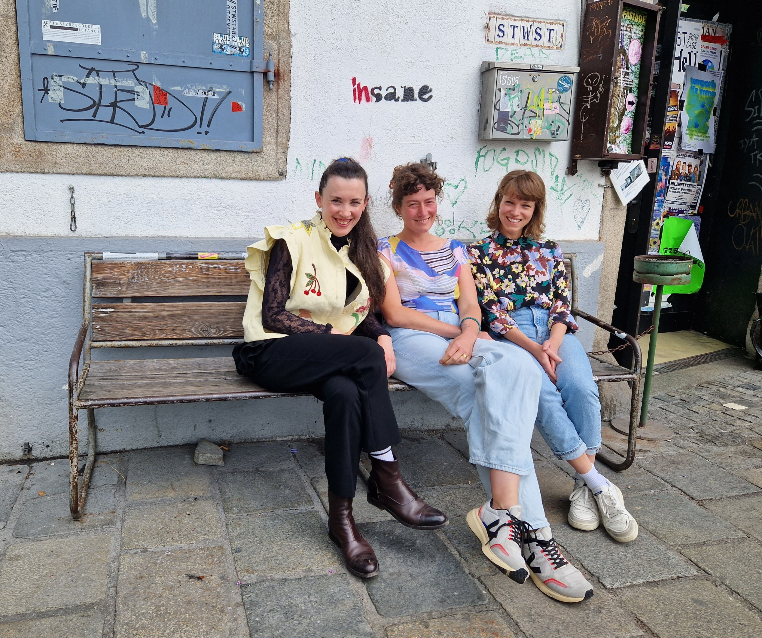 3 Frauen sitzen auf einer Bank vor einem Gebäude und lächeln in die Kamera.