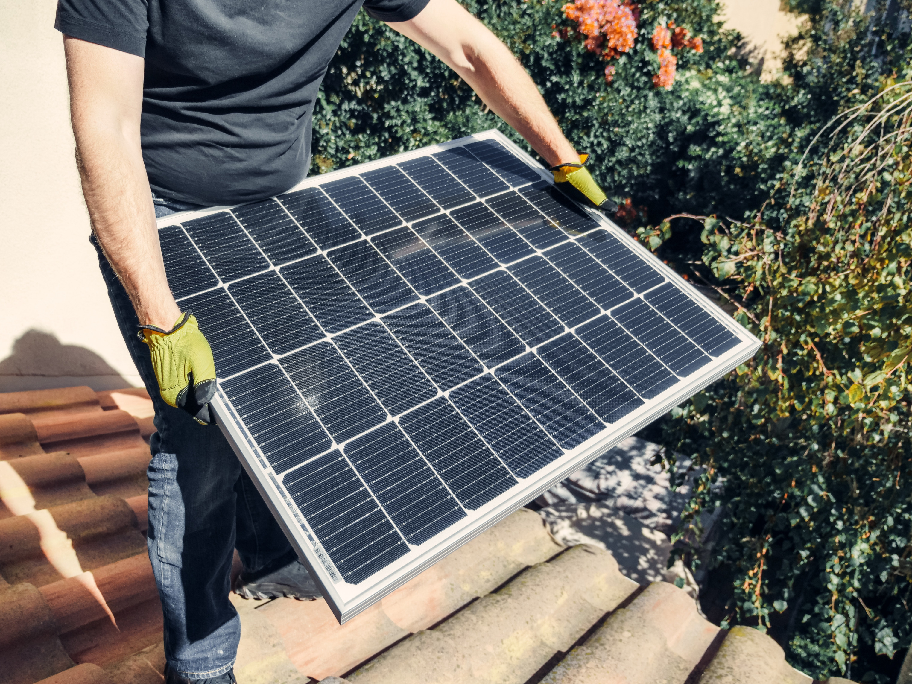 Eine Person hält eine Solarpanel in den Händen. Der Kopf ist nicht zu sehen, die Person trägt gelbe Arbeitshandschuhe