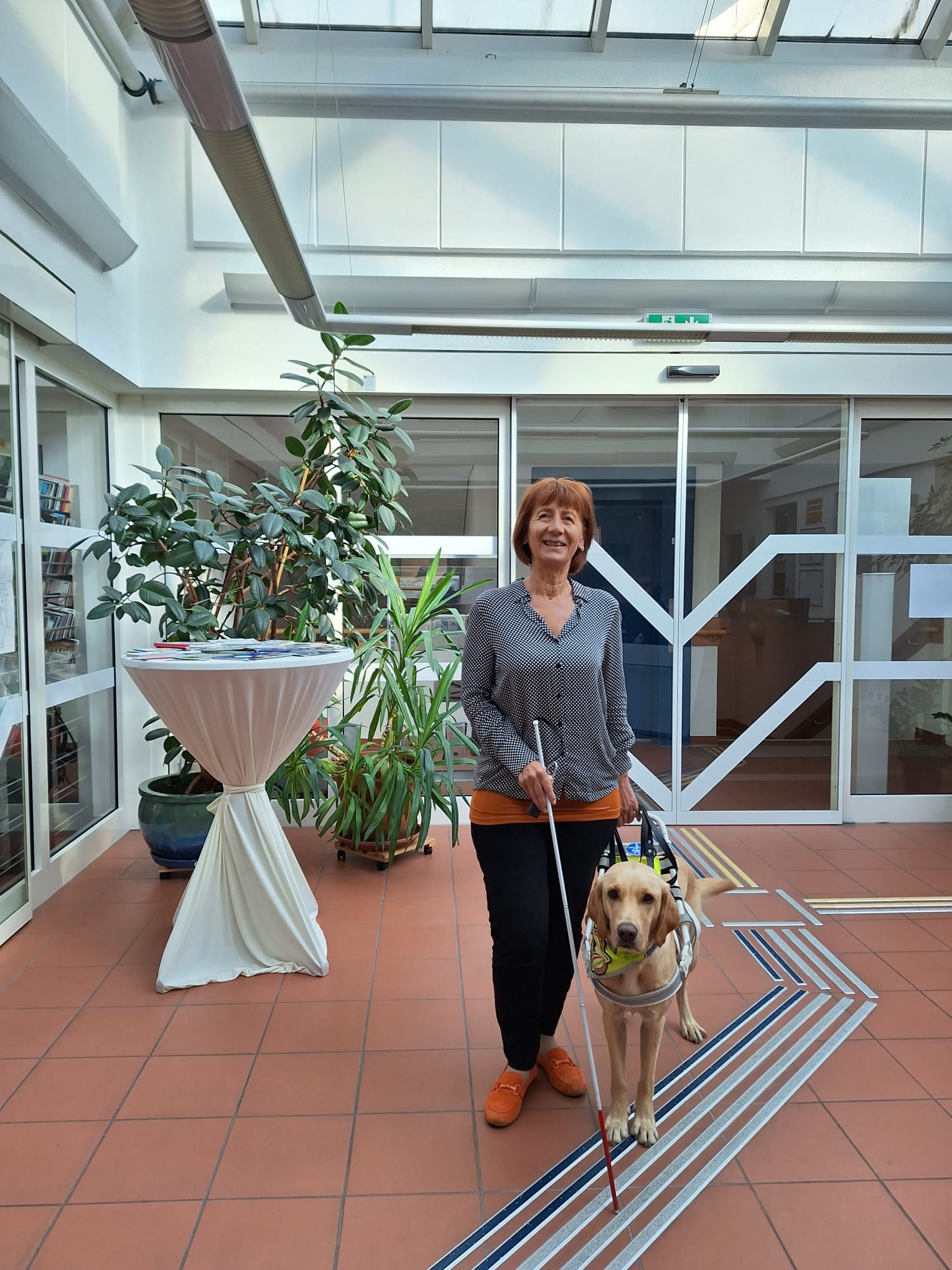 Susanne breitwieser steht mit einem Blindenstock und ihrer Blindenhündin Cora in einem Innenraum. Dieser hat rote Fliesen und ein Blindenleitsystem am Boden. Hinter ihr ist eine Glasschiebetüre zu sehen und Pflanzen.