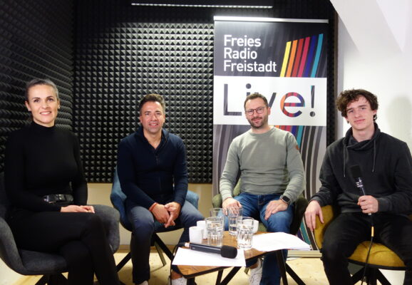 4 Personen sitzen in einem Studio und halten Mikros in den Händen. Sie schauen alle in die Kamera