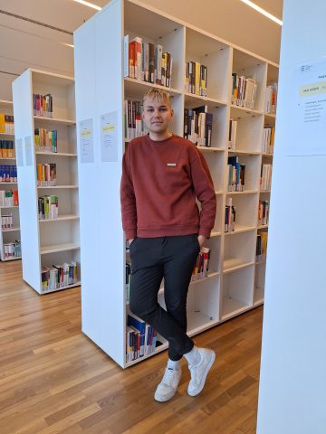 Ein Mann steht gegen ein Bücherregal gelehnt.