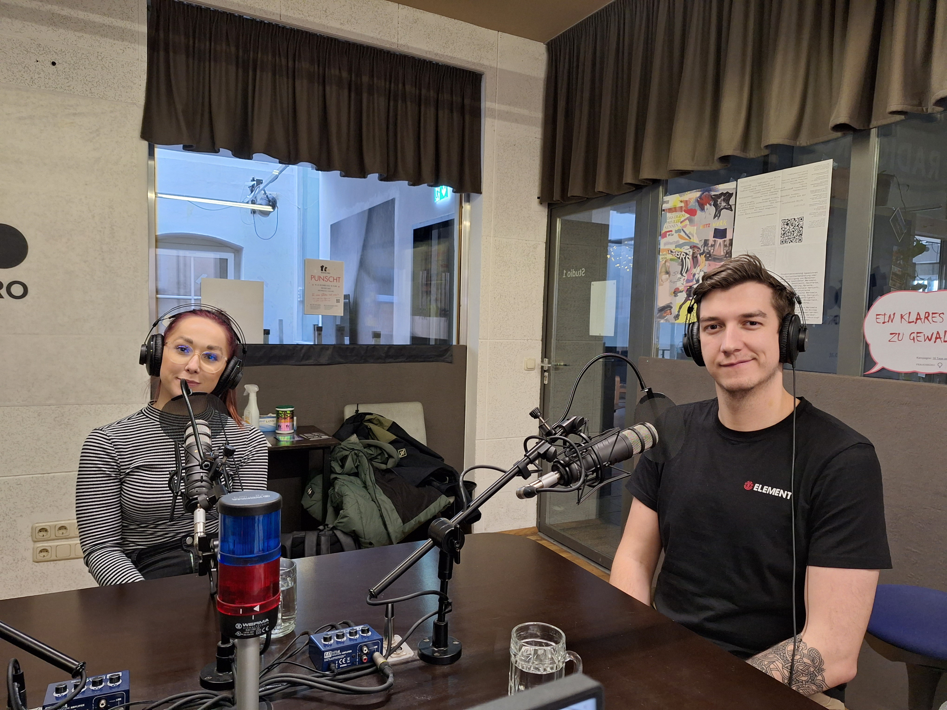 2 Personen sitzen im Radio FRO Studio und schauen in die Kamera. Sie haben Kopfhörer auf.