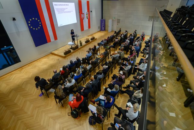 Ein Blick von oben auf einen Saal, in dem Leute in Reihen sitzen. Sie hören einer Frau zu, die auf einem Podium steht und in ein Mikrofon spricht. Hinter ihr wird eine Präsentation auf die Wand projeziert.