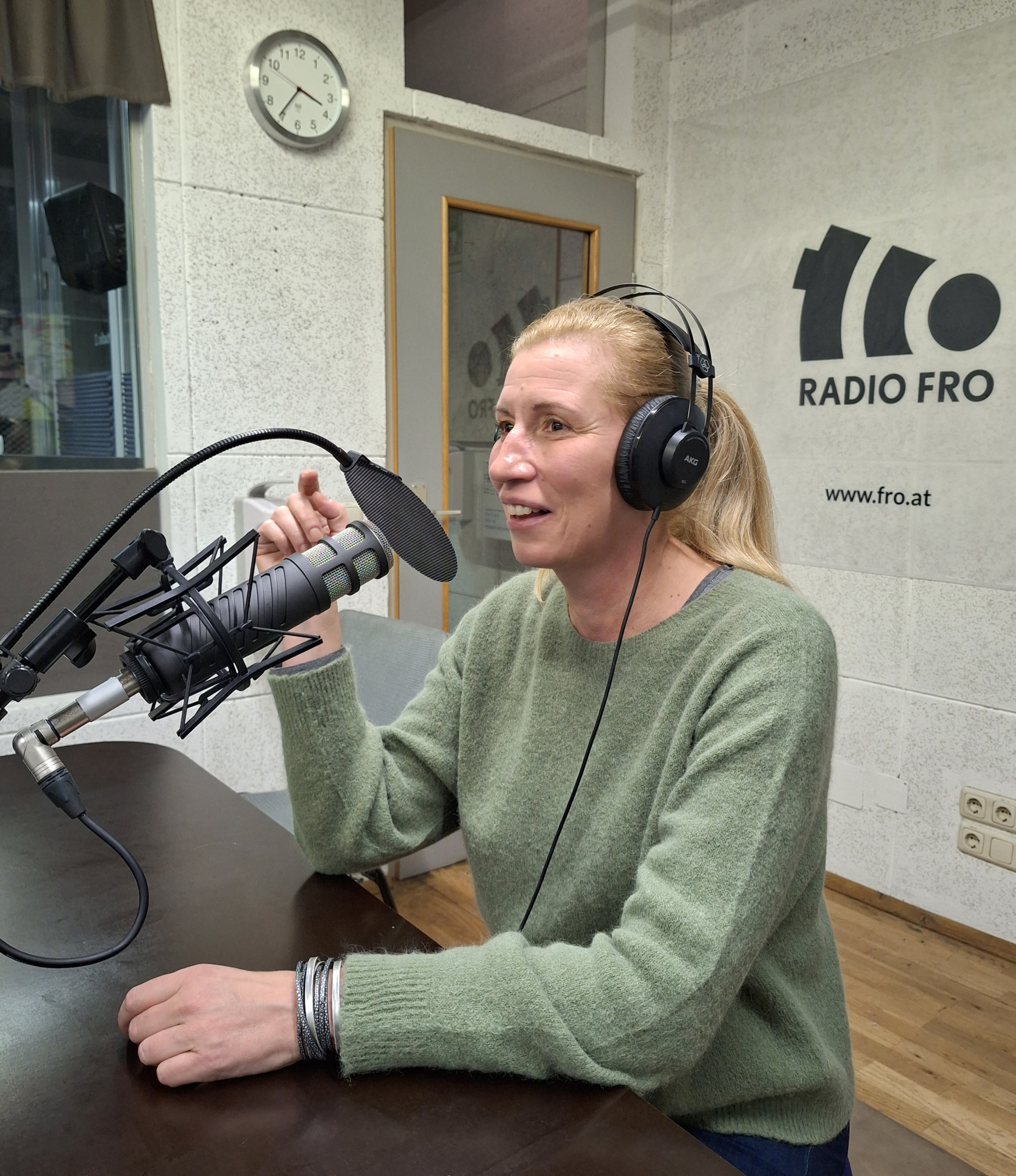 Eine Frau sitzt im Radio FRO Studio. Sie hat Kopfhörer auf und spricht in ein Mikrofon, was vor ihrem Mund steht.