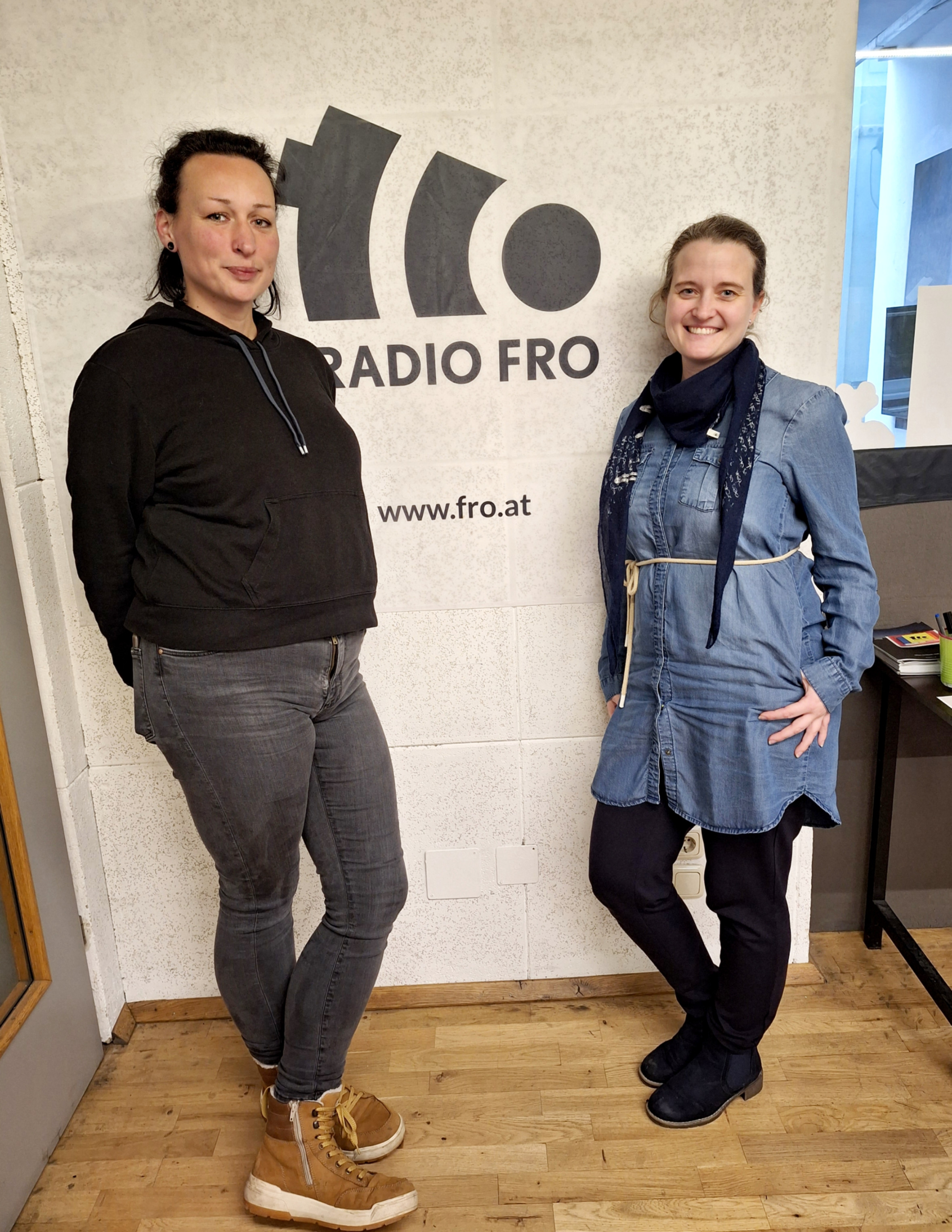 2 Frauen stehen vor einer weißen Wand, auf der das Radio FRO Logo in Schwarz zu sehen ist. 