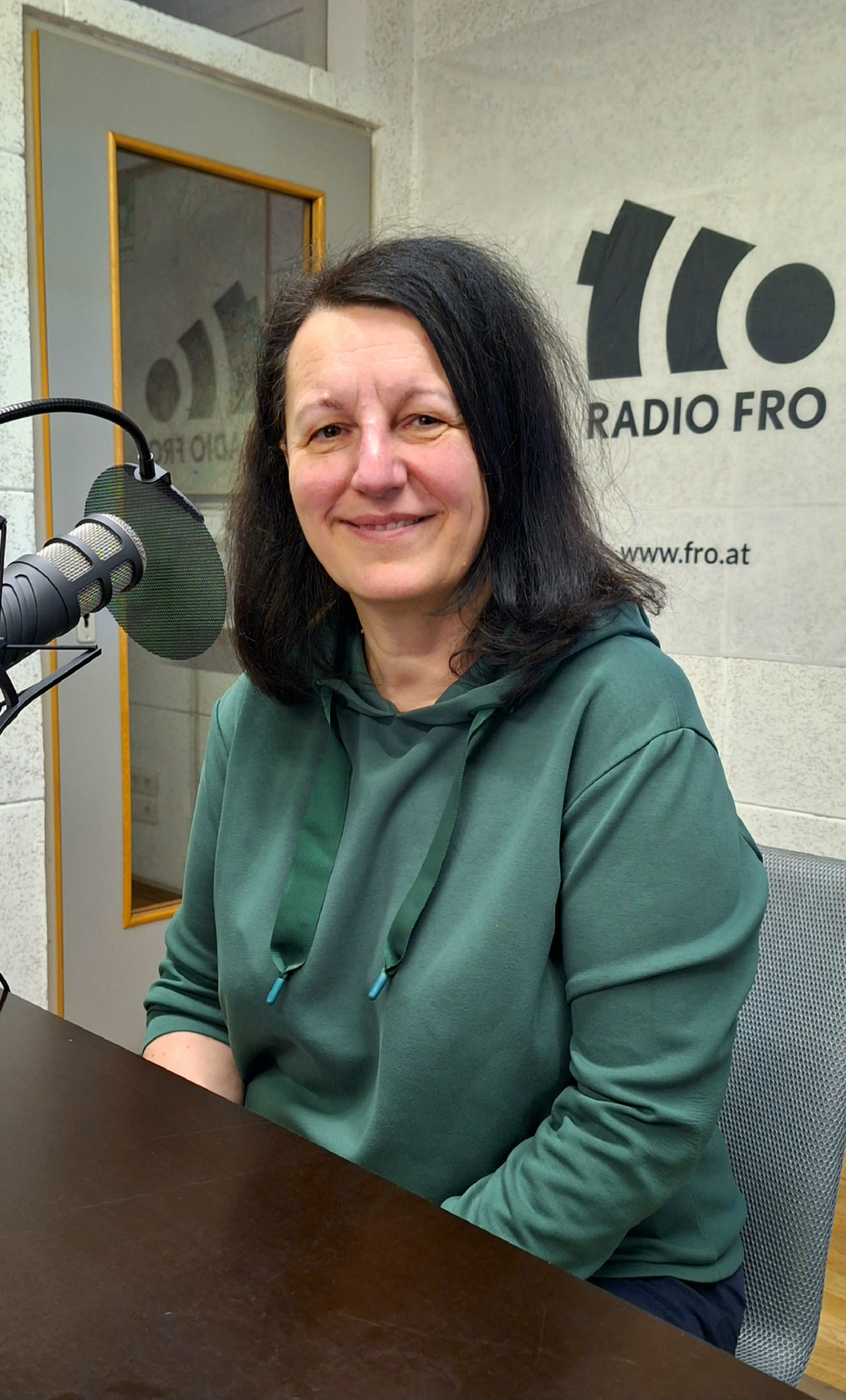 Eine Frau sitzt in einem Radiostudio. Vor ihrem Gesicht ist ein Mikrofon. Sie lächelt in die Kamera