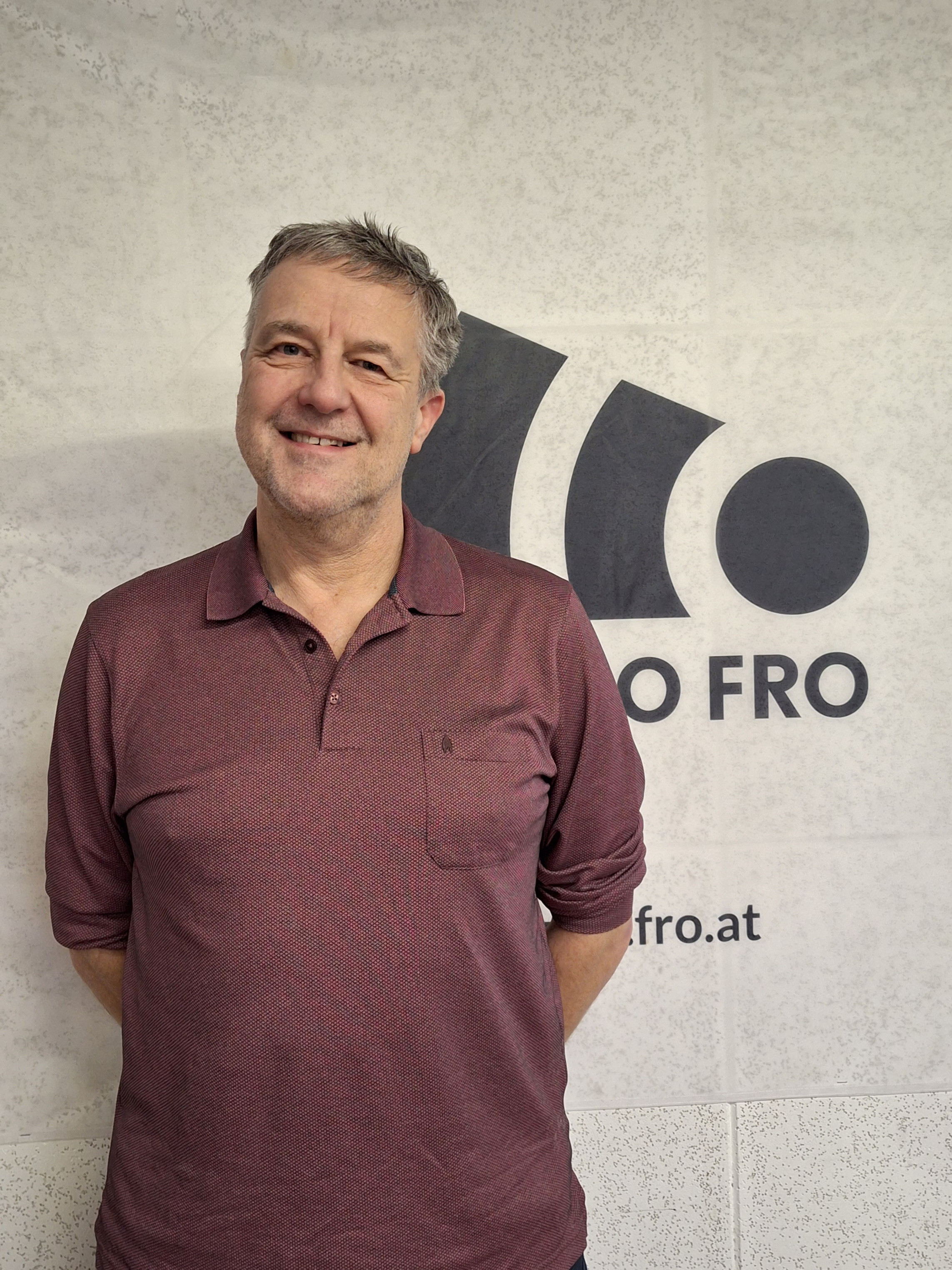 Ein Mann steht lächelnd vor einer weißen Wand auf der das Radio FRO Logo in schwarz zu sehen ist. Er verdeckt das Logo teilweise