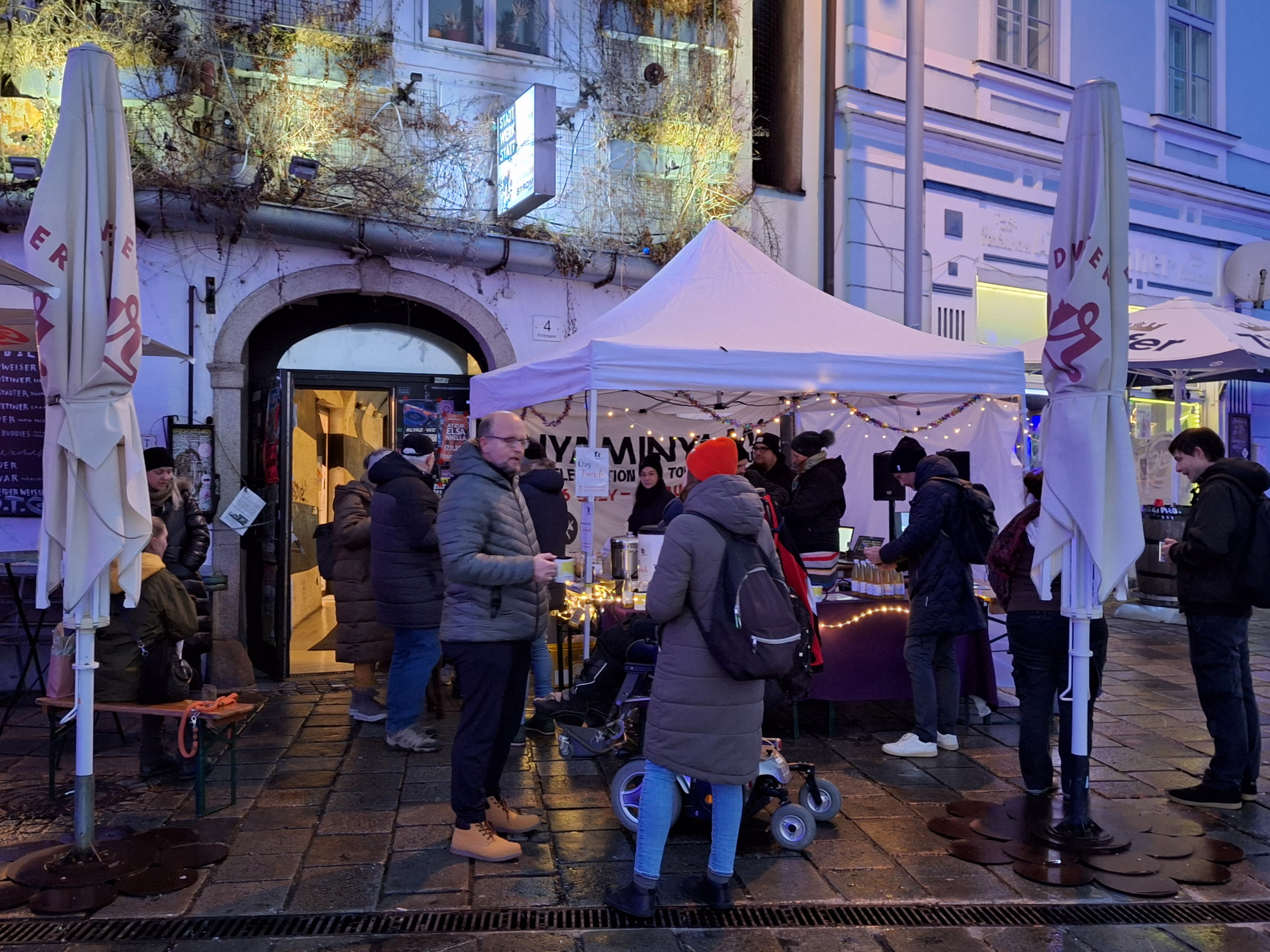 Menschen stehen vor einem Pavillion. Er steht vor der Stadtwerkstatt. Sie reden miteinander und trinken Punsch. Alle haben warme Jacken an.