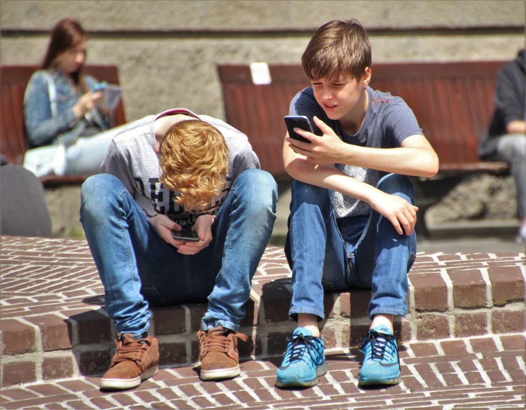 2 Kinder sitzen auf dem Randstein eines Bürgersteigs und schauen in ihre Handys