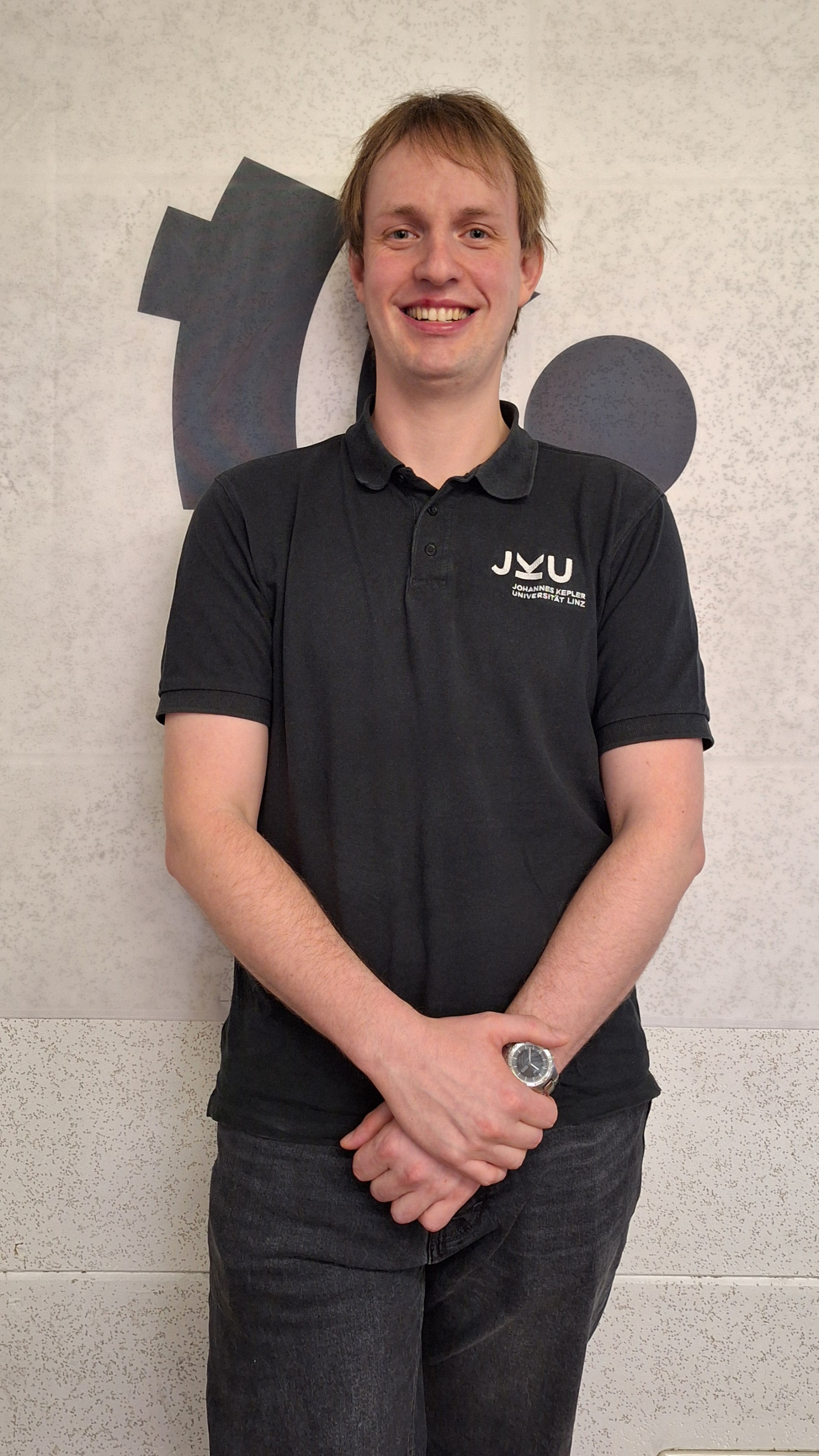 Ein Mann steht vor einer weißen Wand, auf der das Radio FRO Logo zu sehen ist. Seine Hände sind vor dem Körper verschränkt. Er lächelt in die Kamera. Er trägt ein schwartes Poloshirt mit dem Logo der Johannes Keppler Universität