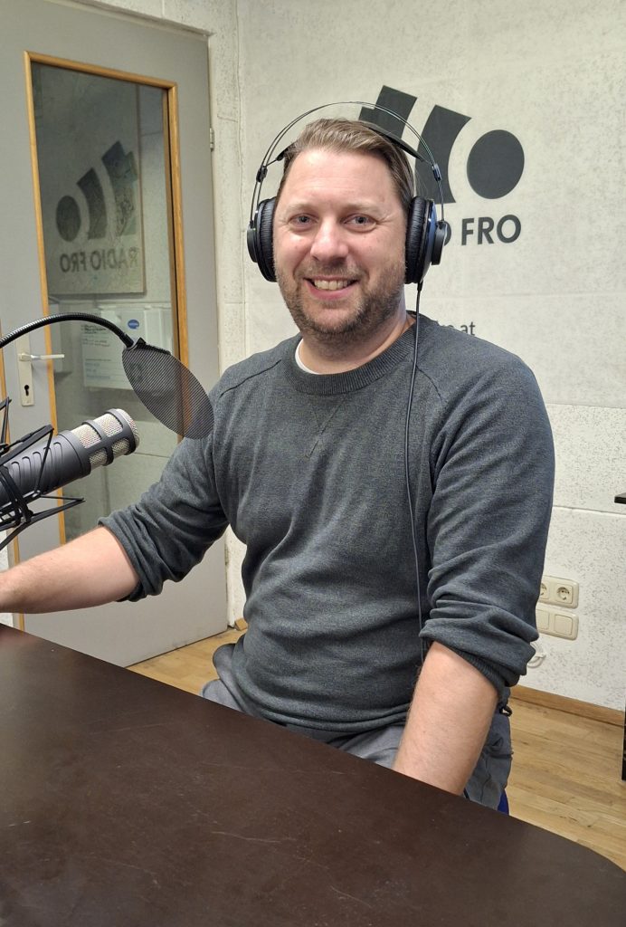 Ein Mann sitzt in einem Radiostudio. Er sitzt an einem Tisch und hat Kopfhörer auf. Links vor ihm ist ein Mikrofon zu sehen. Er lächelt in die Kamera.