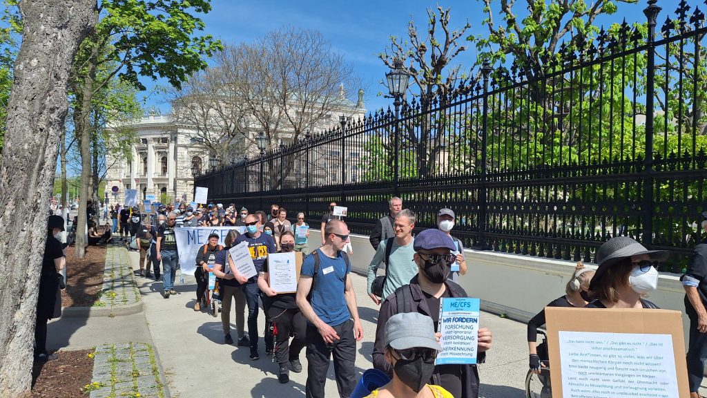 Eine Demo bewegt sich auf die Kamera zu. Viele Menschen haben eine Maske auf und halten Schilder hoch. Es ist sonnig und warm.