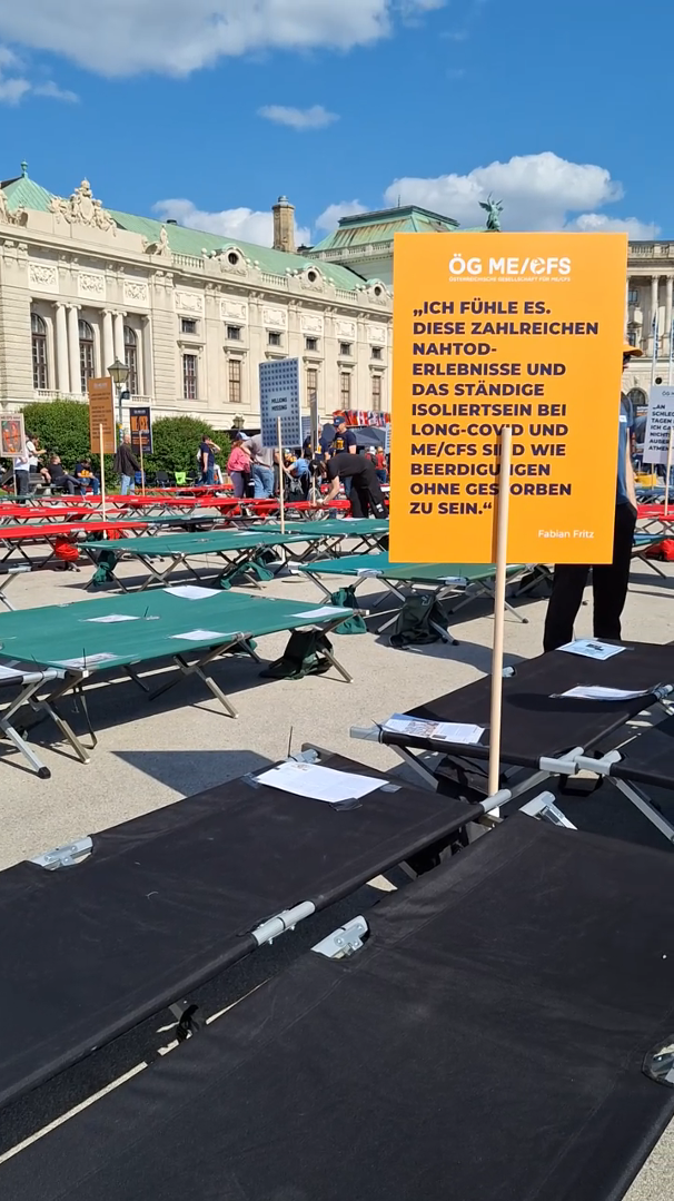 Auf dem Wiener Heldenplatz sieht man Feldbetten in Reihen aufgestellt. Zwischen 2 steckt ein oranges Schild, auf dem steht: "Ich fühle es, diese zahlreichen Nahtoderlebnisse und das ständige isoliertsein bei Long Covid und ME/CFS sind wie Beerdigungen ohne gestorben zu sein."