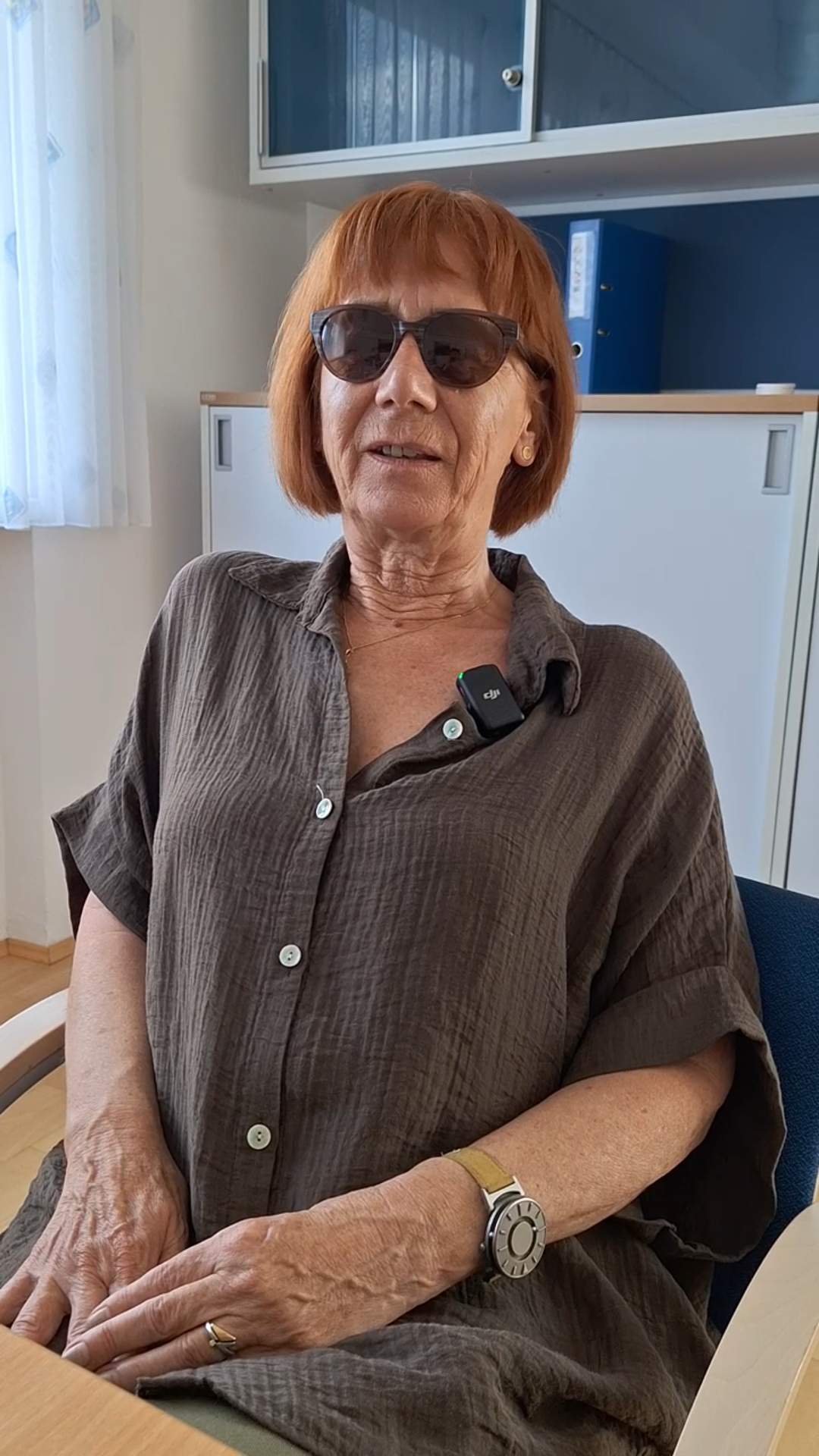 Eine Frau mit kurzen roten Haaren und Sonnenbrille sitzt an einem Holztisch. Sie spricht gerade.