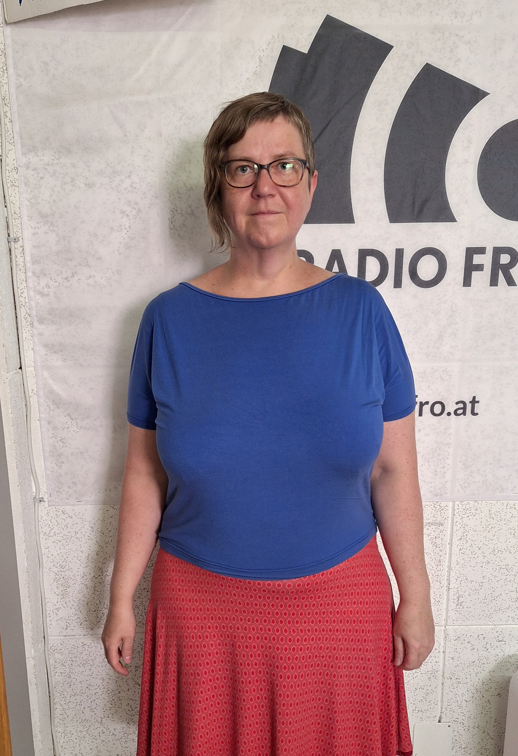 Eine Frau steht vor einer weißen Wand, auf der das Radio FRO Logo in schwarz zu sehen ist. Sie trägt kurze Haare, eine Brille, ein blaues Shirt und einen roten Rock.