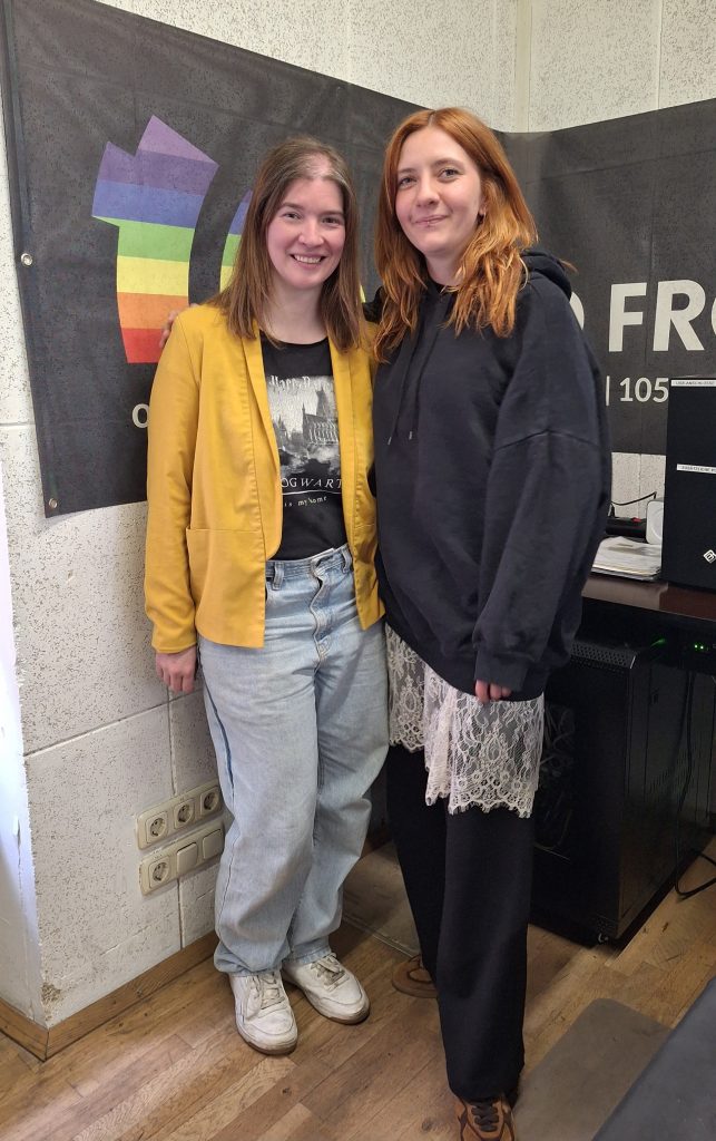 Zwei Frauen stehen vor dem Radio FRO Logo Banner in einem Radiostudio. Beide lächeln in die Kamera