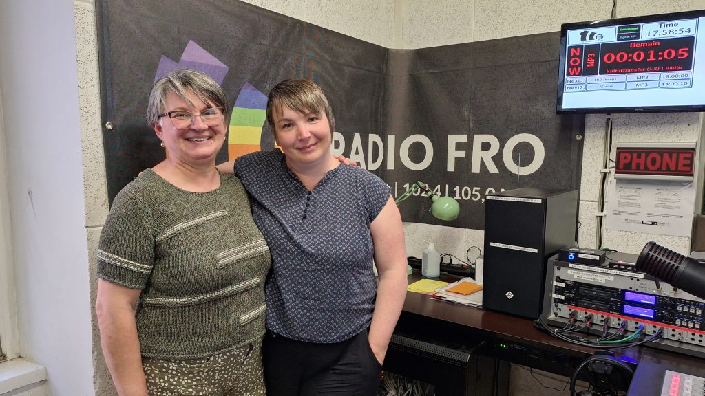 2 Frauen stehen in einem Radiostudio. Sie haben die Arme gegenseitig über die Schultern gelegt und lächeln in die Kamera. Hinter ihnen hängt ein schwarzes Banner mit dem Radio FRO Logo an der Wand. 